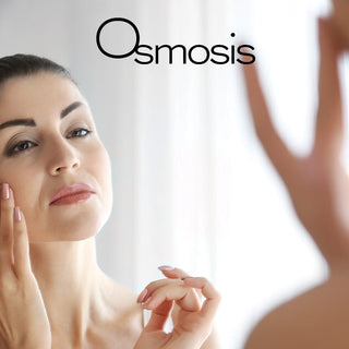 Woman examining her clear healthy skin in mirror while using Osmosis skincare products.
